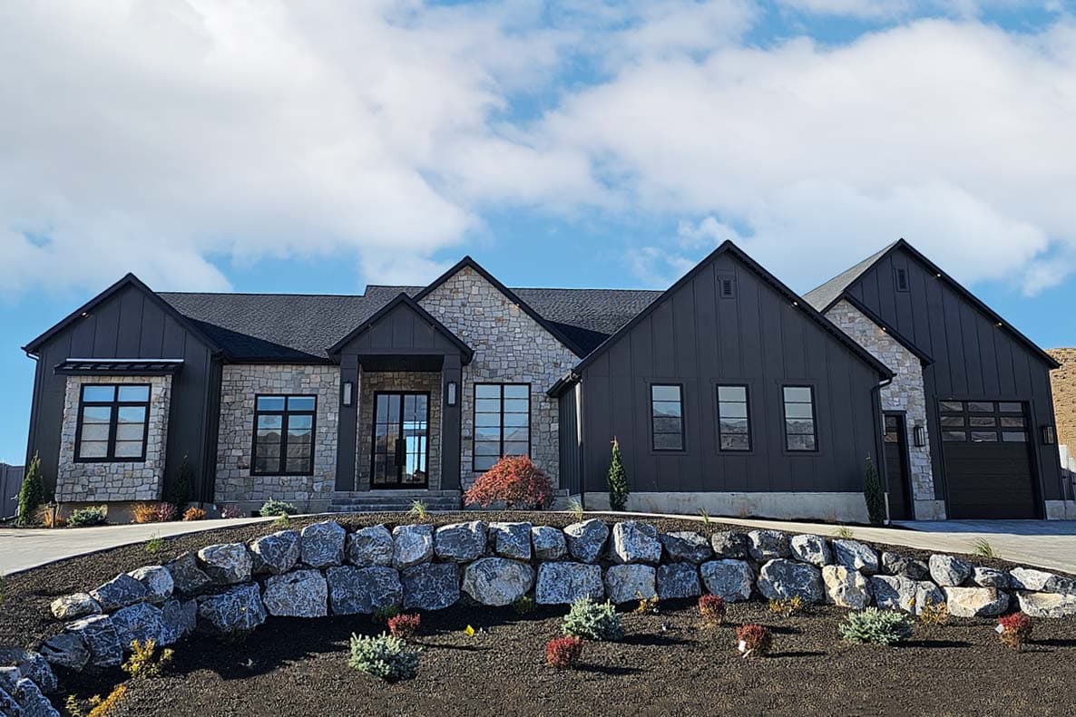 House plan exterior. Modern Farmhouse style with two gabled sections, stone and dark siding, and a two-car garage. Stone retaining wall in foreground.