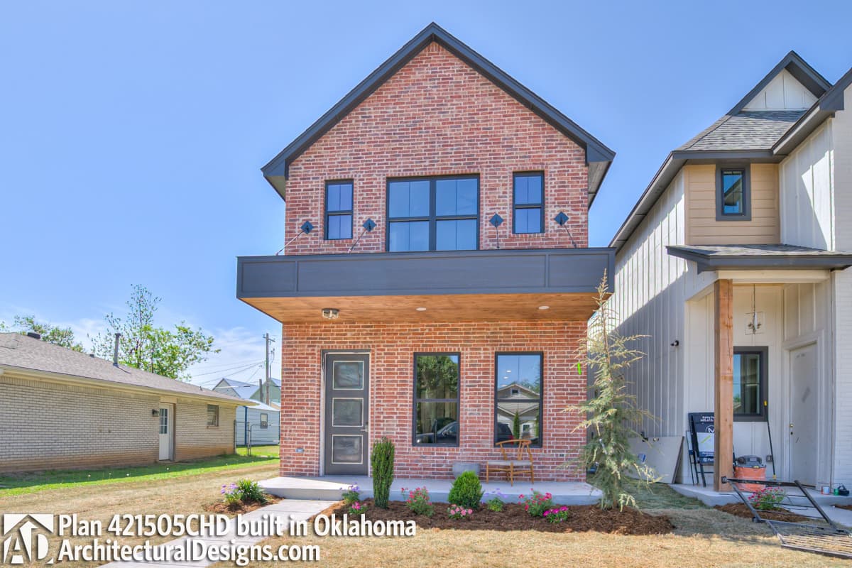 Two-story house plan exterior with brick facade, dark gables, and cantilevered second-story balcony.