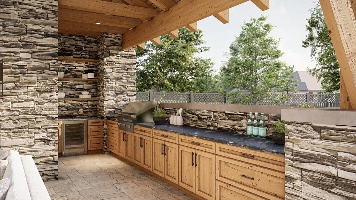 Outdoor kitchen with stone walls and wooden cabinets. Grill and countertop are set against a backdrop of trees and a fence.