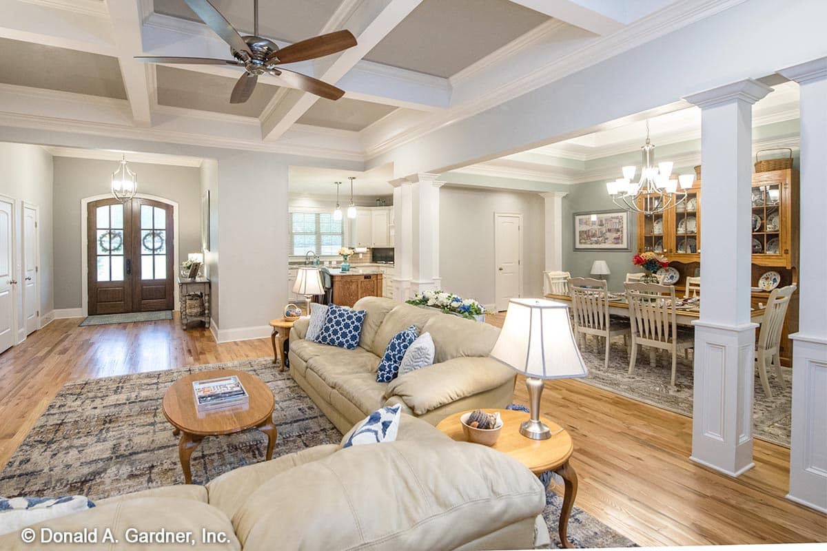 Interior view of a house plan showing a living room with coffered ceiling, double front doors, and adjoining dining room and kitchen.