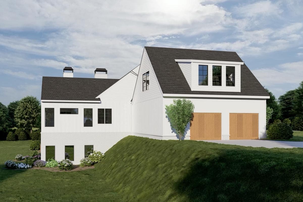 A modern white house with a dark roof and wooden garage doors sits atop a small grassy hill. Lush green trees surround the home.
