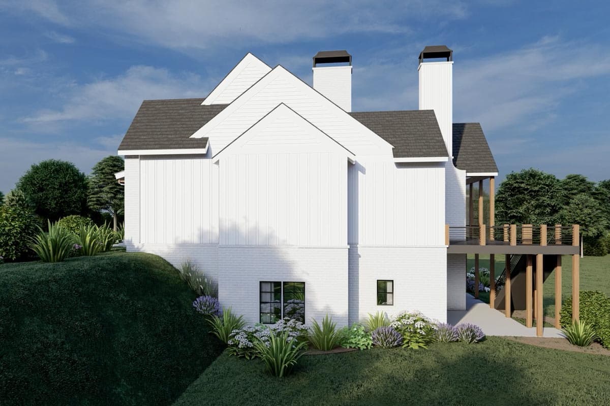 Side view of a modern white house with a gabled roof, two chimneys, and a wooden deck. Landscaping includes bushes and flowers on a grassy hill.