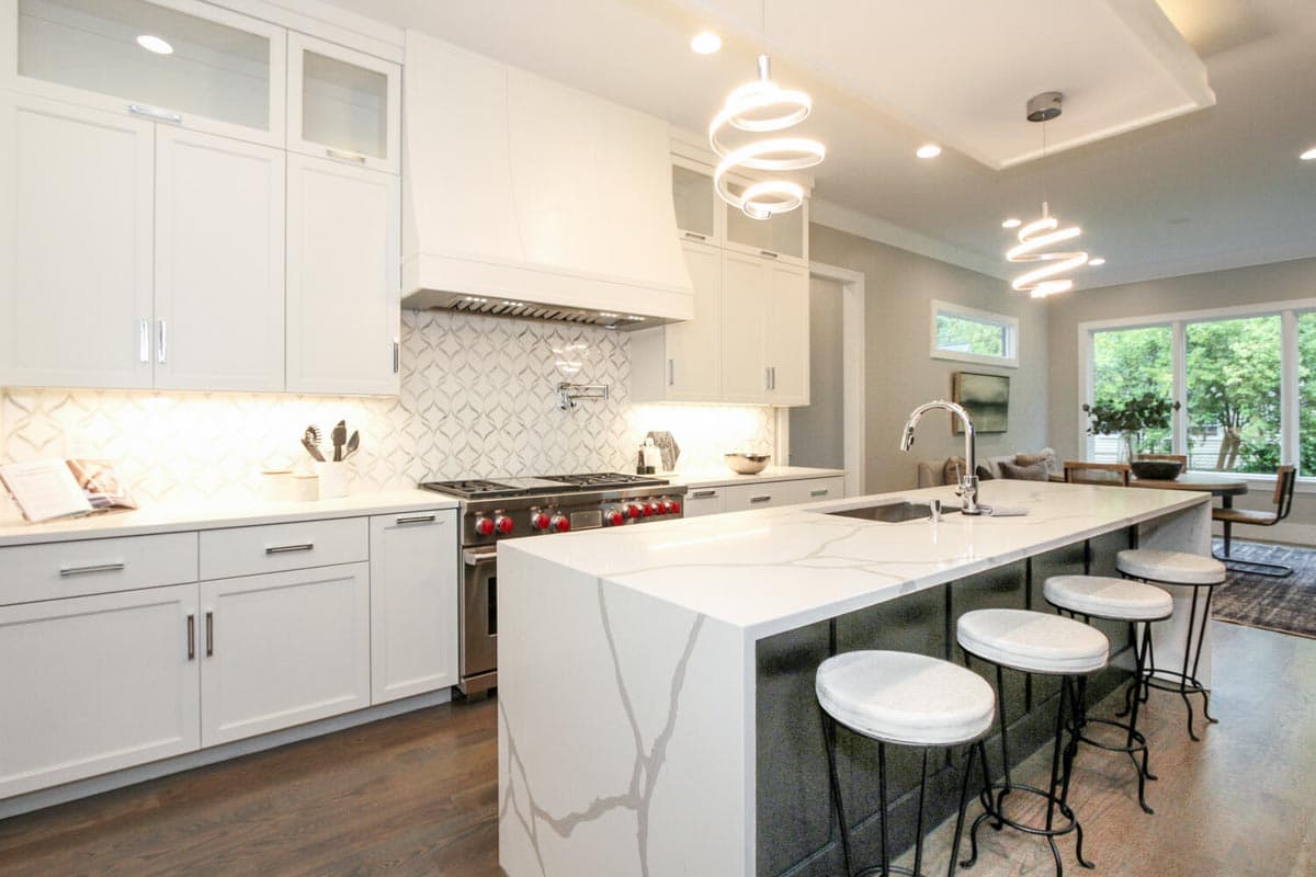 Modern kitchen with white cabinets, marble island with sink, stainless steel range, and pendant lighting. Open to dining area.