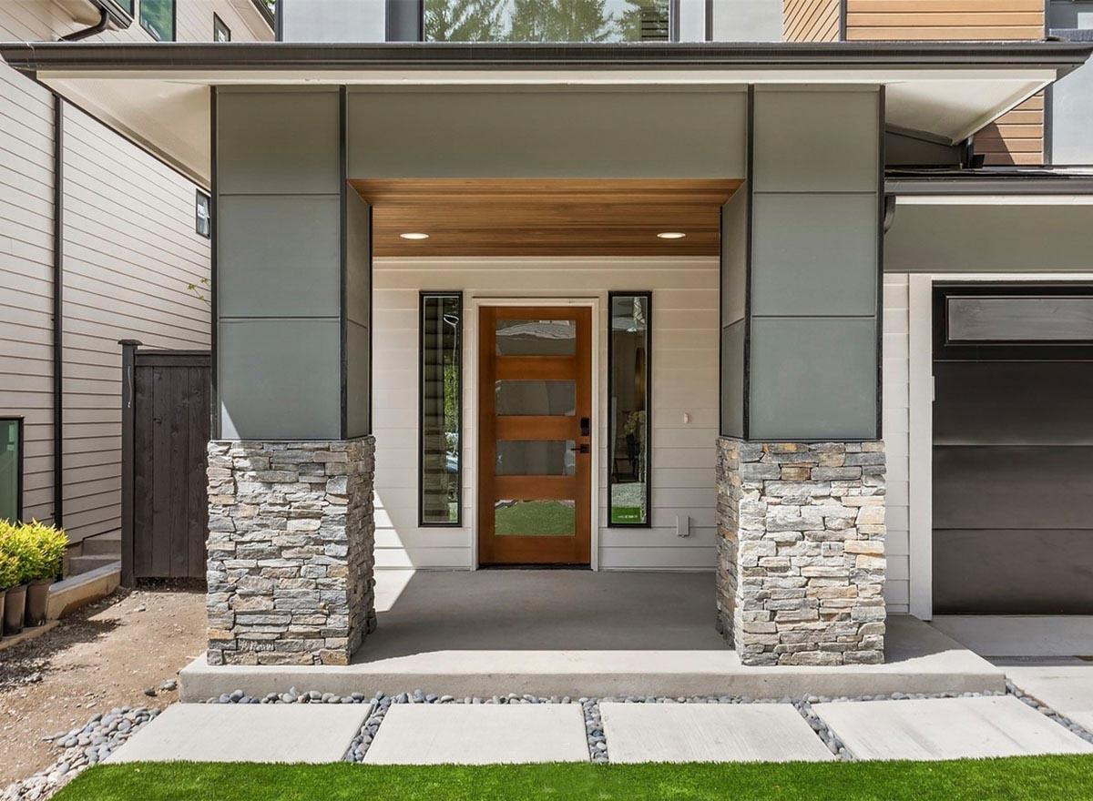 Modern house exterior with stone-clad columns, wood-paneled ceiling, and a frosted glass pane entry door.