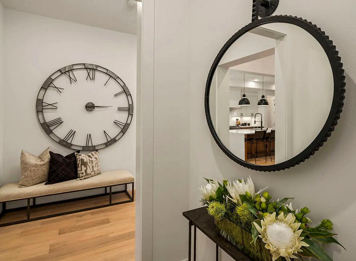 Interior view of a hallway with large Roman numeral clock, bench, and reflection of kitchen with island and pendant lights.