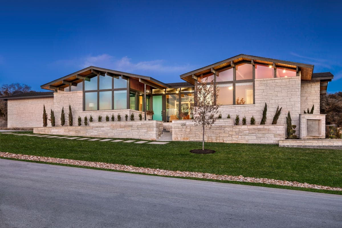 House plan exterior. Modern style, single story. Features include large windows, exposed beams, and a stone facade. Entry has a turquoise door.