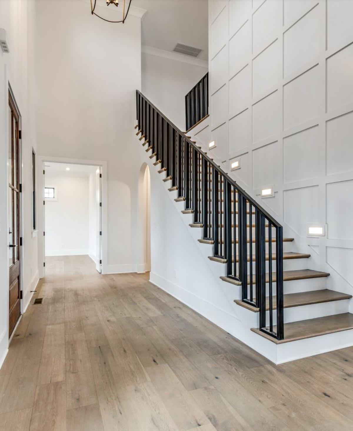 Interior hallway with open staircase, wood flooring, and paneled wall with sconces.
