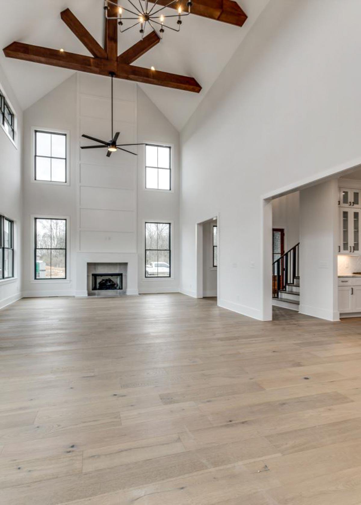 Great room with vaulted ceiling, exposed wood beams, fireplace, and large windows. Opens to kitchen and staircase.