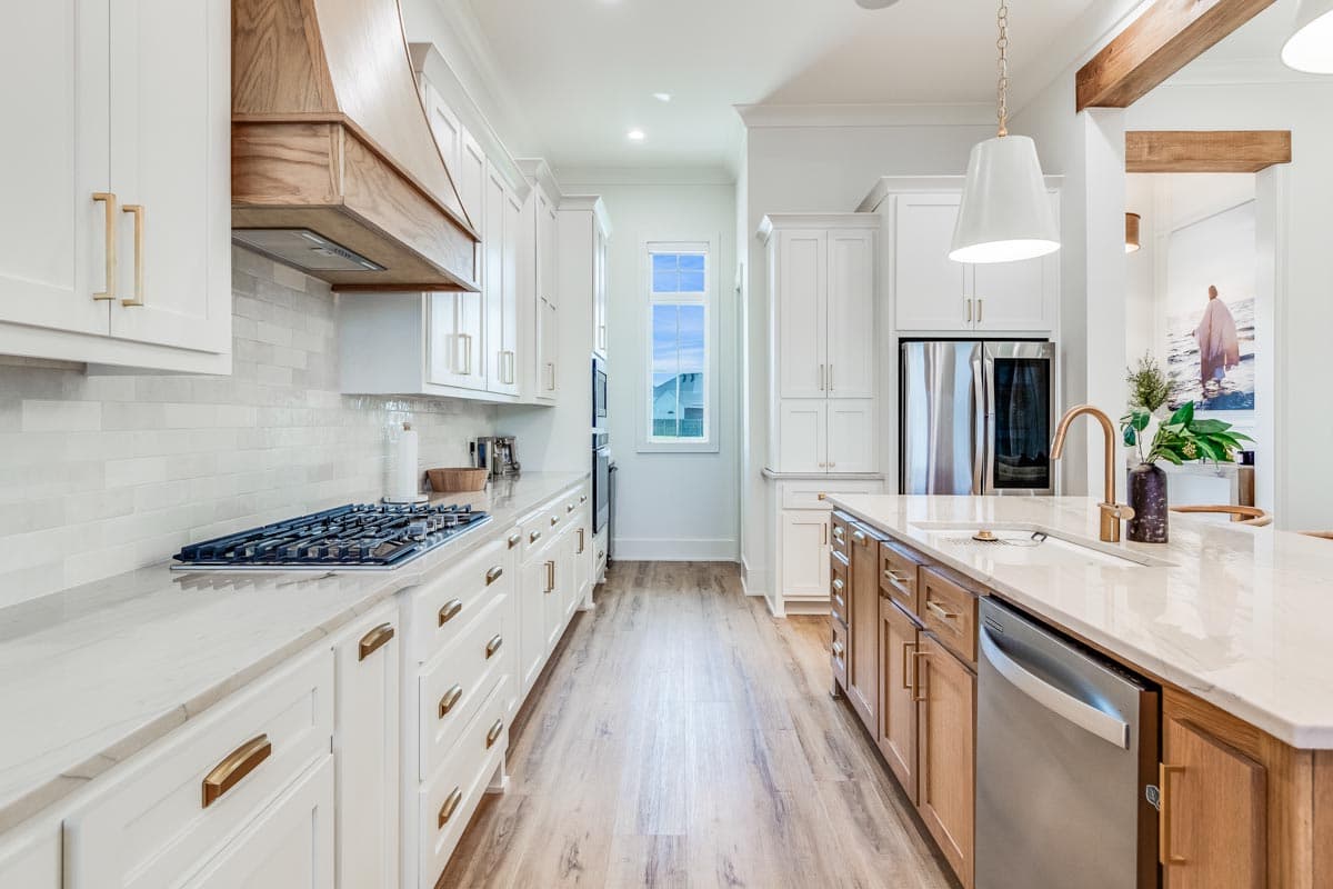 Kitchen interior with white cabinetry, wood range hood, and center island. Features include gas cooktop, pendant lighting, and stainless steel appliances.