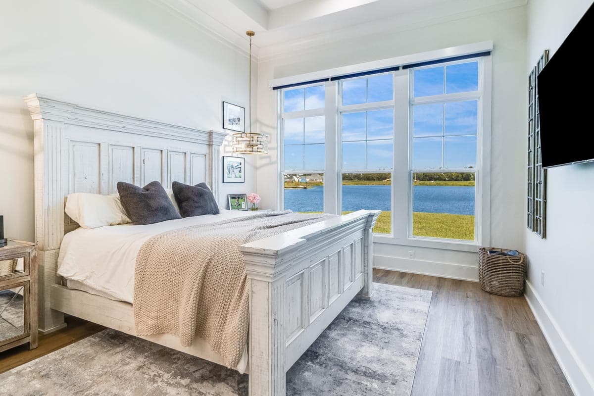 Primary suite interior with large windows overlooking a lake. Features include a white bed, decorative light fixture, and molding.
