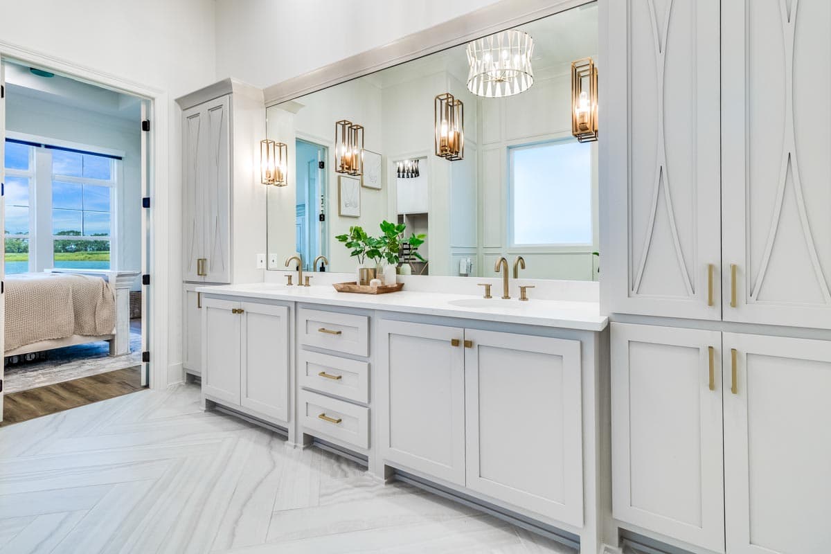 Bathroom interior with double vanity, large mirror, decorative lighting, and paneled cabinetry. Open doorway leads to bedroom.