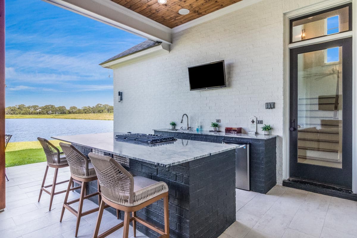 Outdoor kitchen with a brick island, bar seating, built-in appliances, a mounted TV, and access to a door with glass panels.