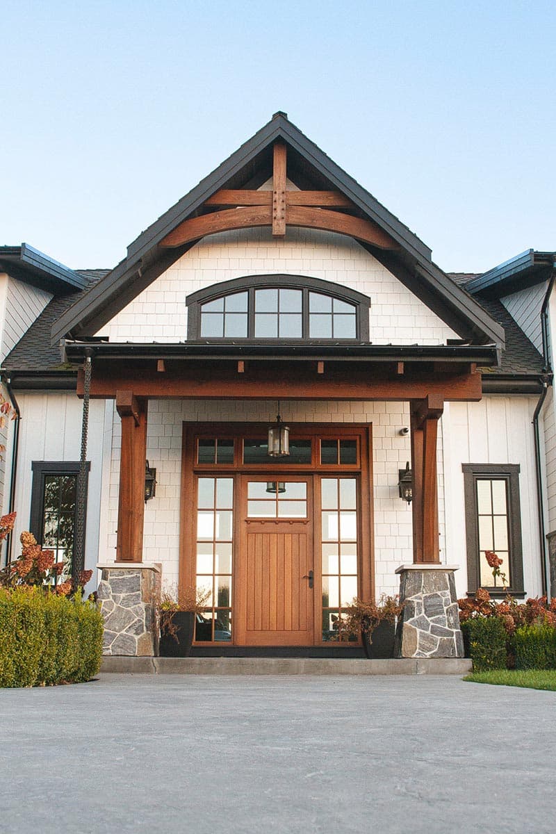 Modern farmhouse exterior with a timber-framed gable, wood front door, stone column bases, and a transom window.