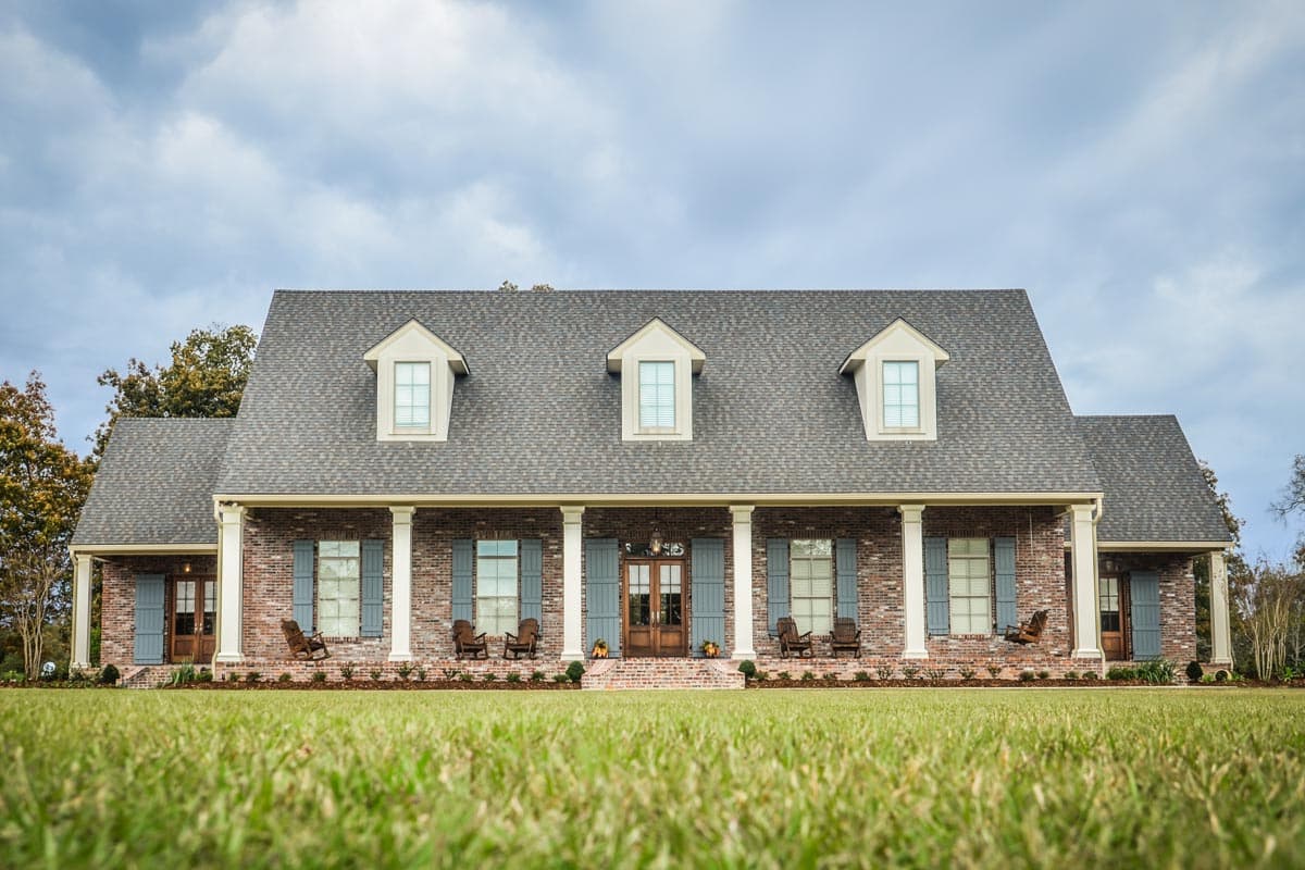 House plan exterior. Two-story brick home with dormers, a covered porch, and columns. Includes shutters and a gabled roof.