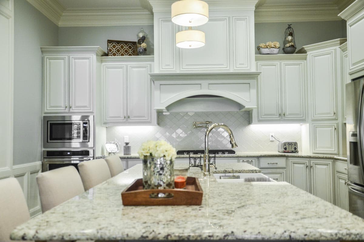 Kitchen with white shaker cabinets, granite island, arched range hood, mosaic tile backsplash, and pendant lighting.