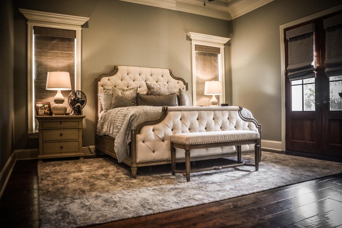 Bedroom interior with tufted king bed, upholstered bench, nightstand with lamp, and double wood doors with glass panes.