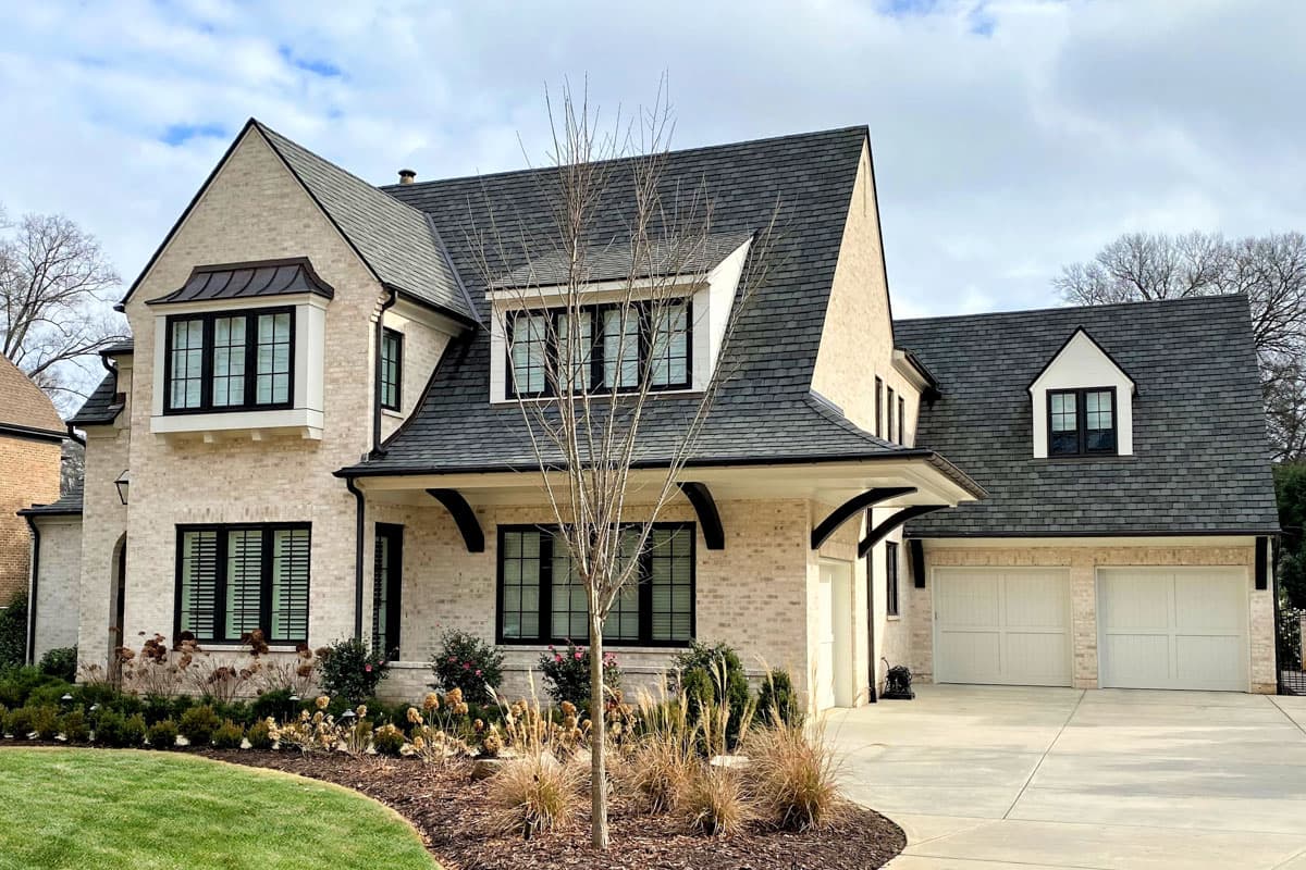 House plan exterior: light brick two-story residence with steeply pitched roof, multiple gables, bay window, and attached two-car garage.