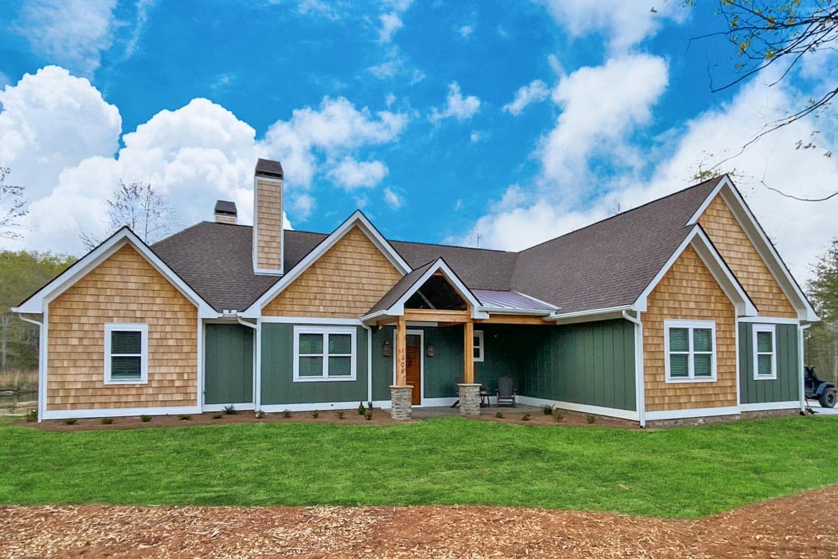 A craftsman-style house with green siding and cedar shake accents sits under a partly cloudy sky. The home has a welcoming porch and lush green lawn.