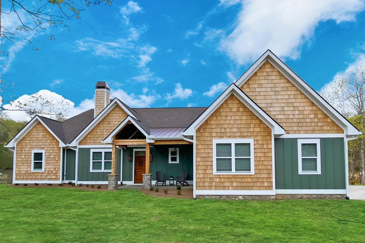A craftsman-style house with green siding, cedar shake shingles, and a welcoming front porch. The house is on a grassy lawn with a blue sky.