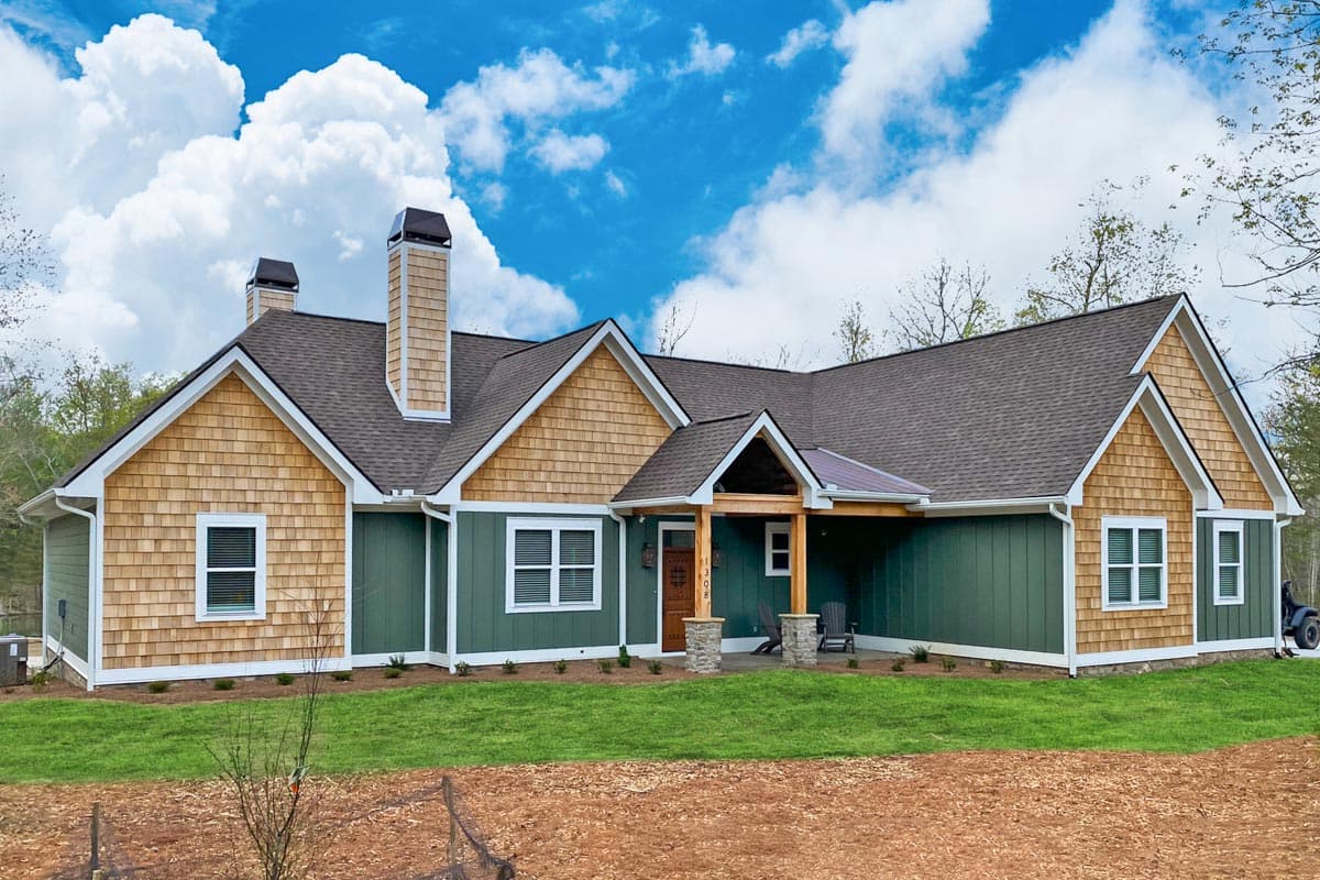 A craftsman-style house with green siding, cedar shake accents, and a covered porch. Lush green lawn contrasts against the blue sky and fluffy clouds.