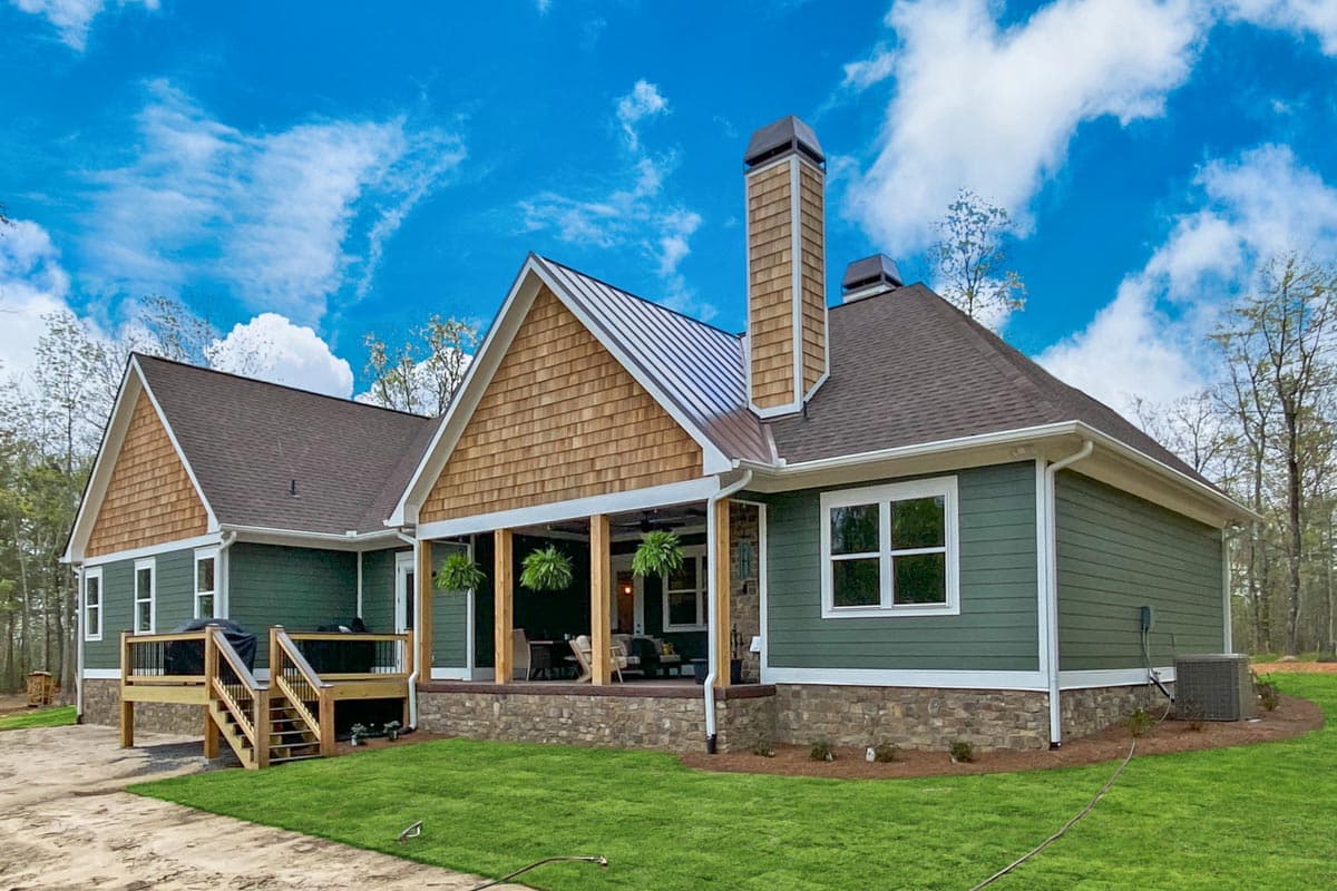 A modern, green-sided house with a stone base, wooden deck, and a metal roof against a bright blue sky with fluffy clouds.