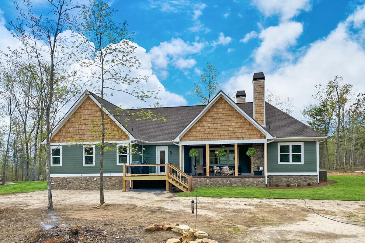 A newly constructed house with green siding, wooden shingle accents, and a covered porch. Lush grass borders the home, against a bright blue sky.