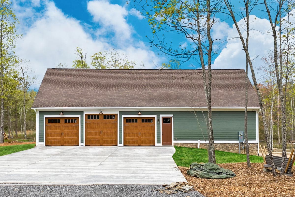 A three-car garage with brown doors sits in front of a blue sky. The building is green with a brown roof and is surrounded by trees.