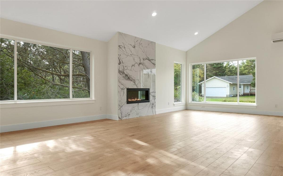 Open concept living space with vaulted ceiling, gas fireplace, and large windows overlooking trees and a garage.