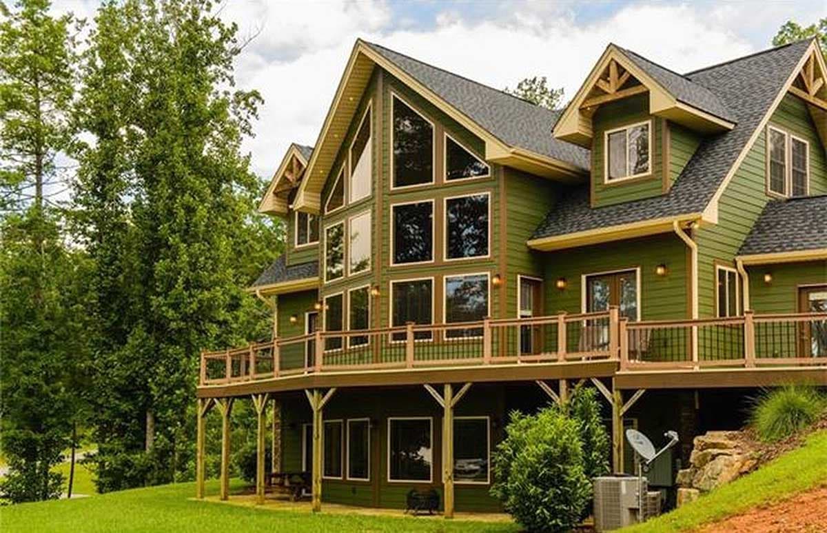 House plan exterior: green two-story home with large multi-pane windows, prominent gables, and a wrap-around deck.