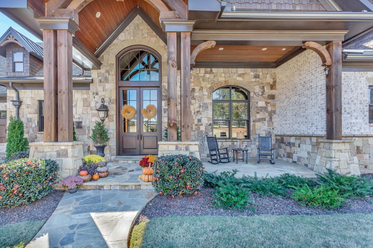 House plan exterior featuring a covered porch with large stone pillars, arched double doors, stone and brick facade, and multiple gables.
