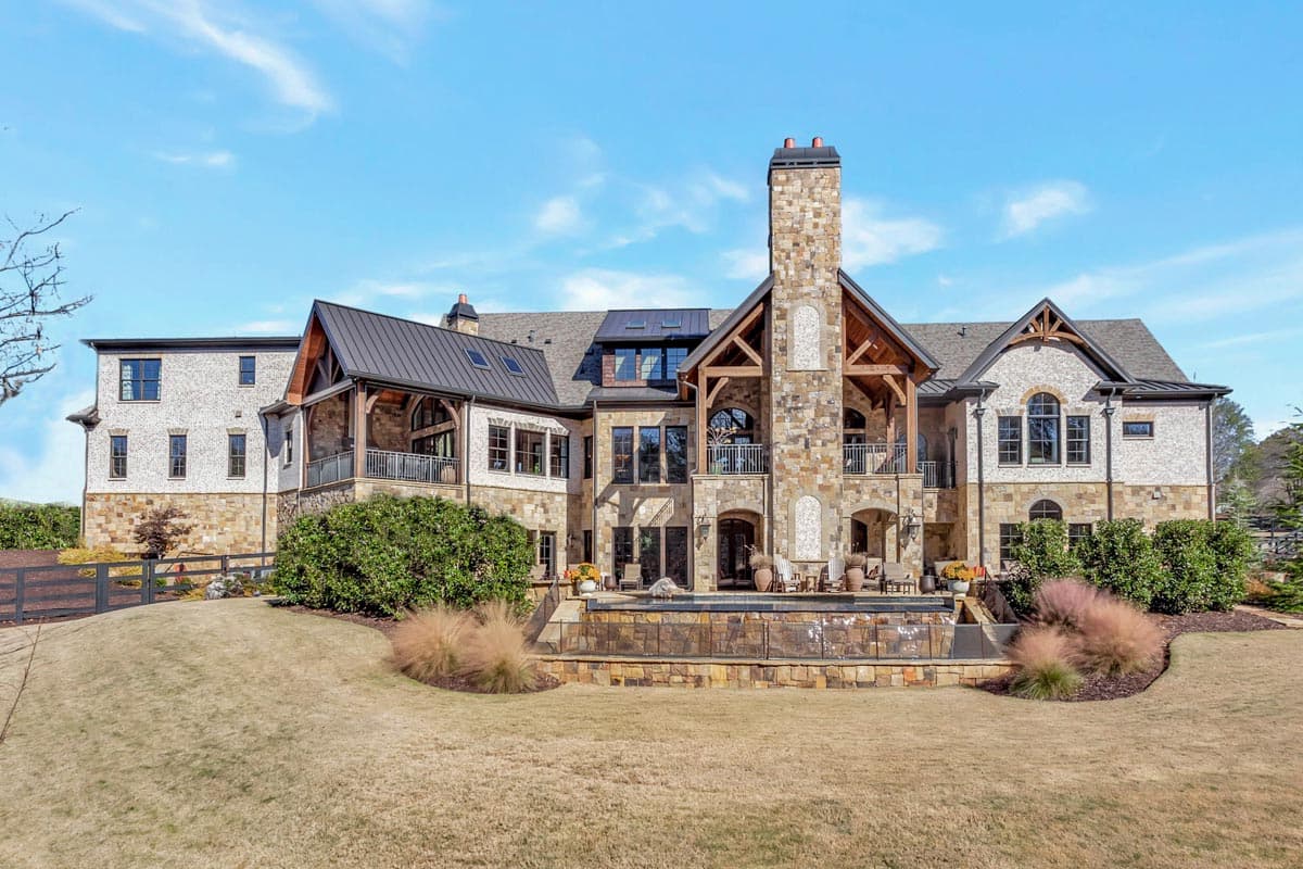 House plan exterior with stone and brick facade, multiple gables, covered balconies, and prominent stone chimney.