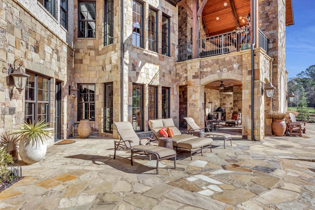 Exterior of a multi-story house with stone facade, covered porch with arched entrance, and large patio with chaise lounges.