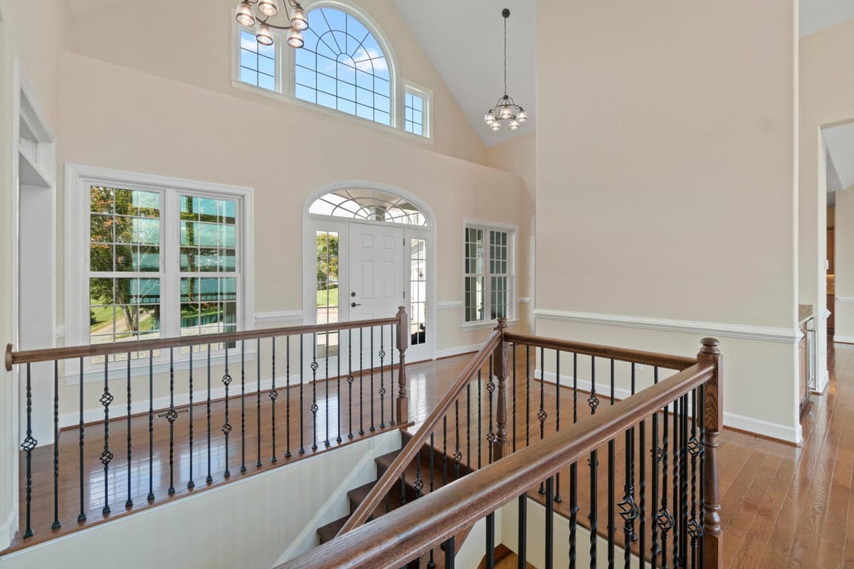 Interior view of a house foyer with a grand staircase, vaulted ceiling, large arched window, and double front doors.