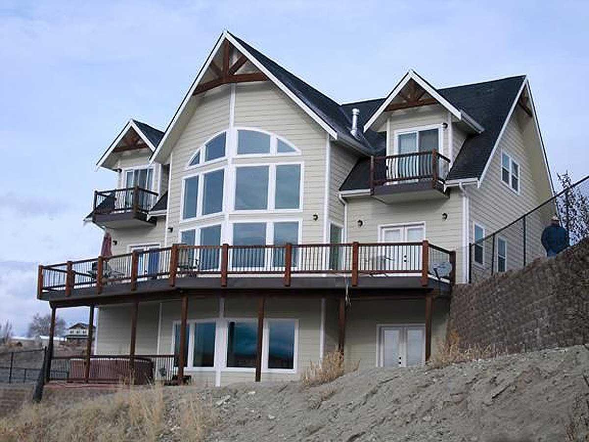 House plan exterior: two-story home with multi-pane gable dormers, wraparound deck, and a large arched window feature.