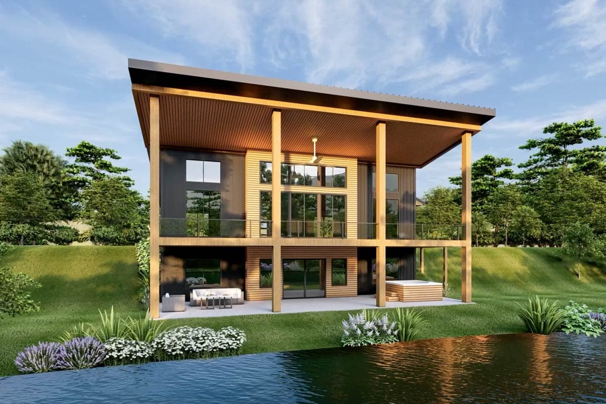A modern two-story home with large windows and a wrap-around deck. It has a distinctive roof design, surrounded by a lake and lush greenery.