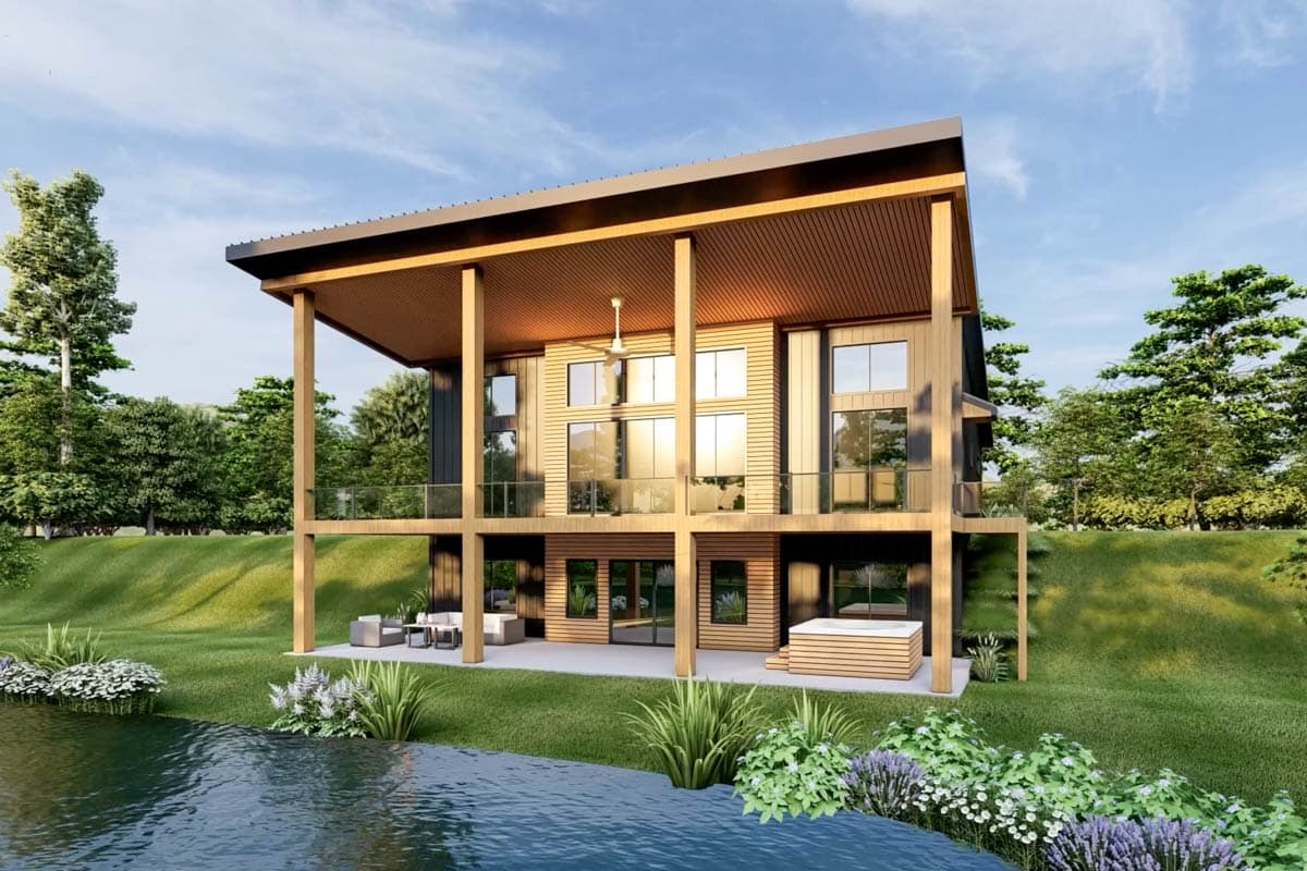 A modern two-story home with large windows and a covered deck, near a lake. Supported by wooden pillars, with a hot tub and seating area.
