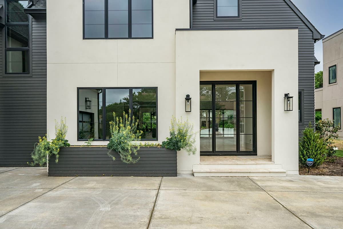 Modern house exterior with stucco and dark siding, featuring large black-framed windows and double glass doors.