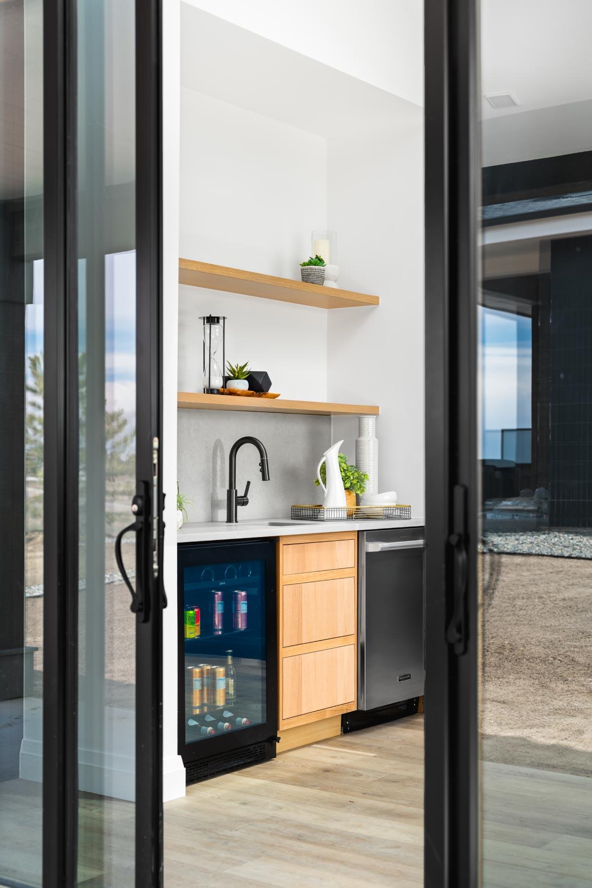 Wet bar with beverage cooler, stainless steel dishwasher, light wood cabinetry, open shelving, and black faucet.