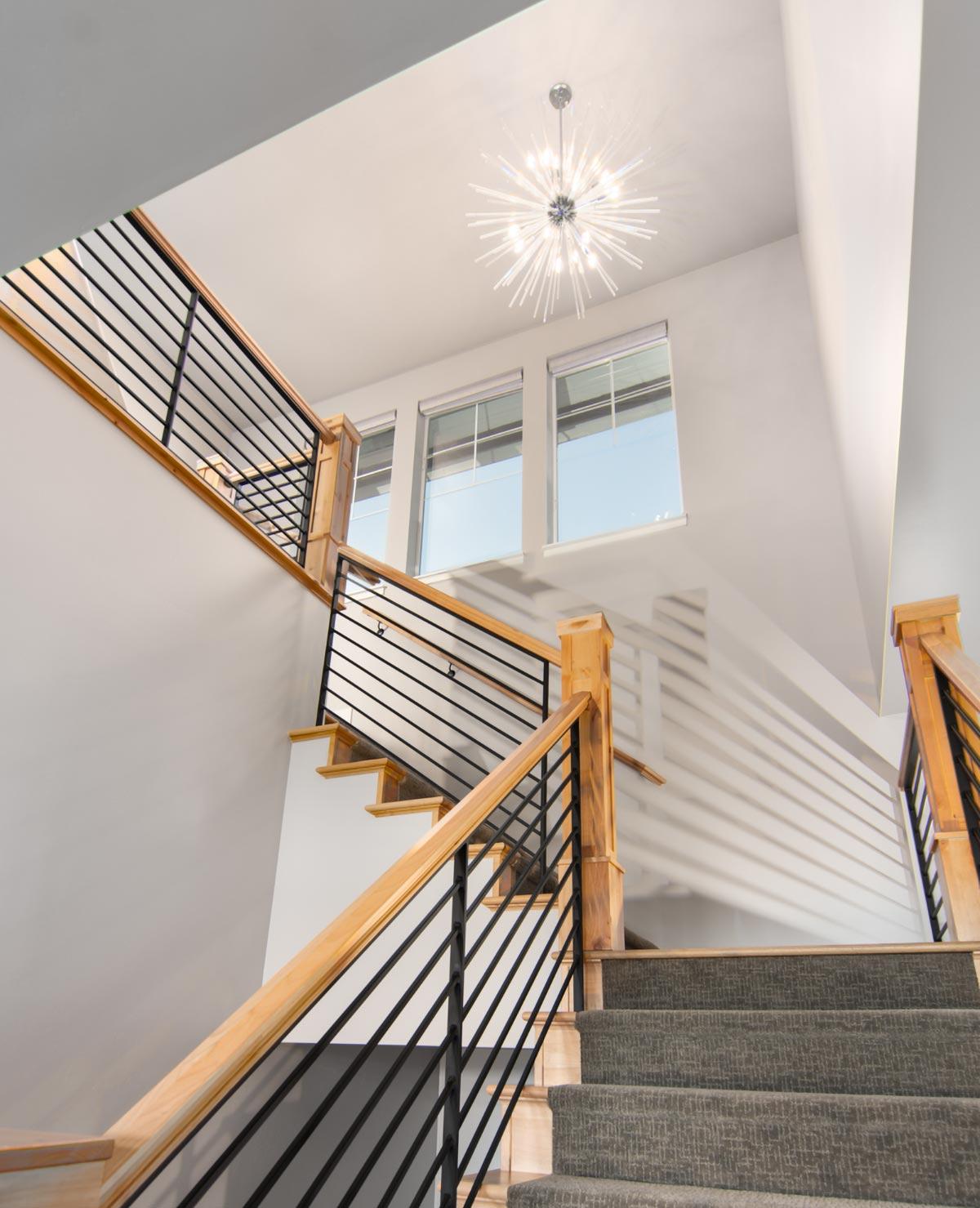 Interior view of a staircase with natural wood posts, black metal railings, carpeted steps, and a large chandelier overhead.