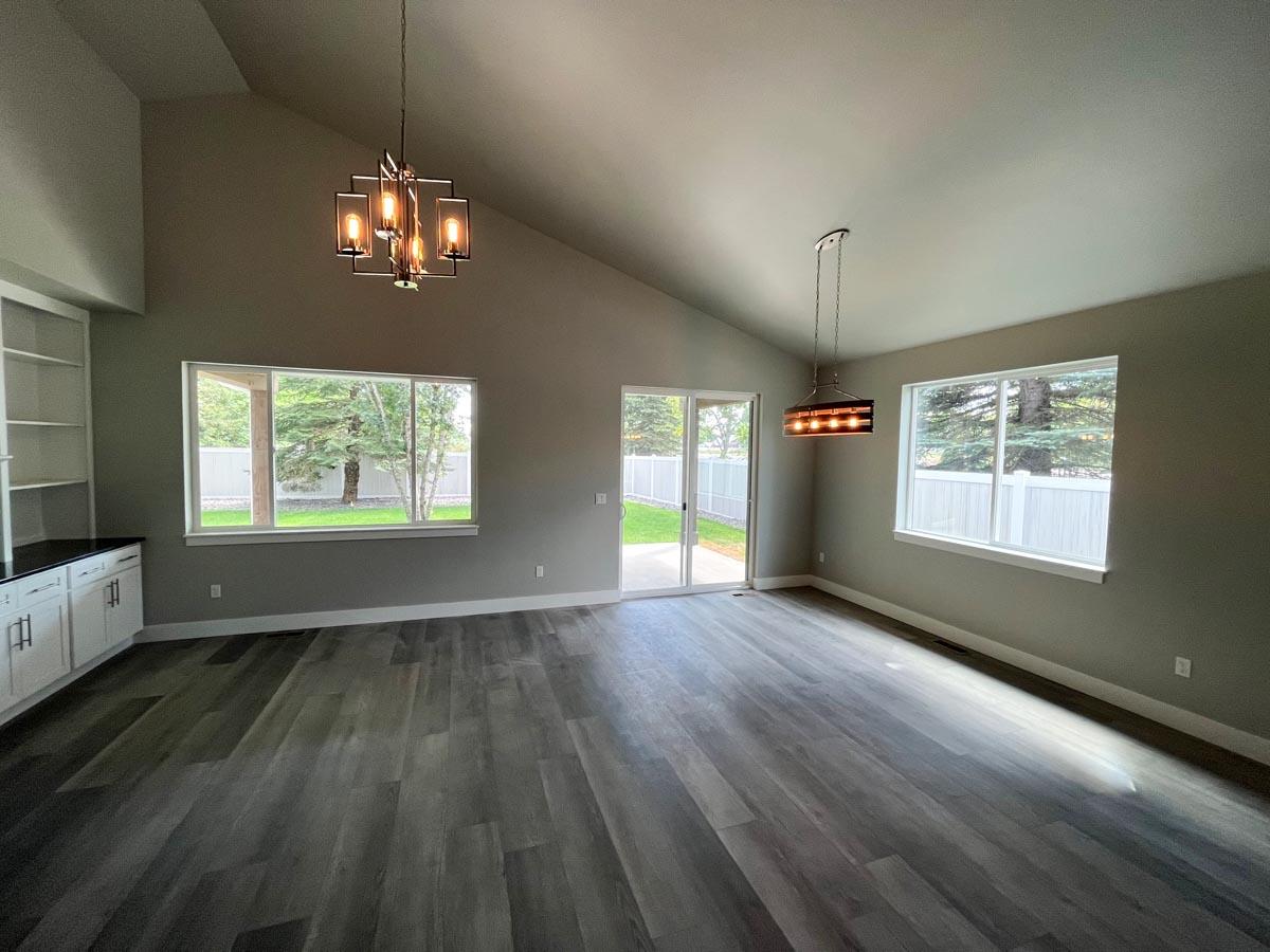 Open-concept interior with vaulted ceiling, large windows, two modern chandeliers, built-in shelving, and sliding glass doors to yard.