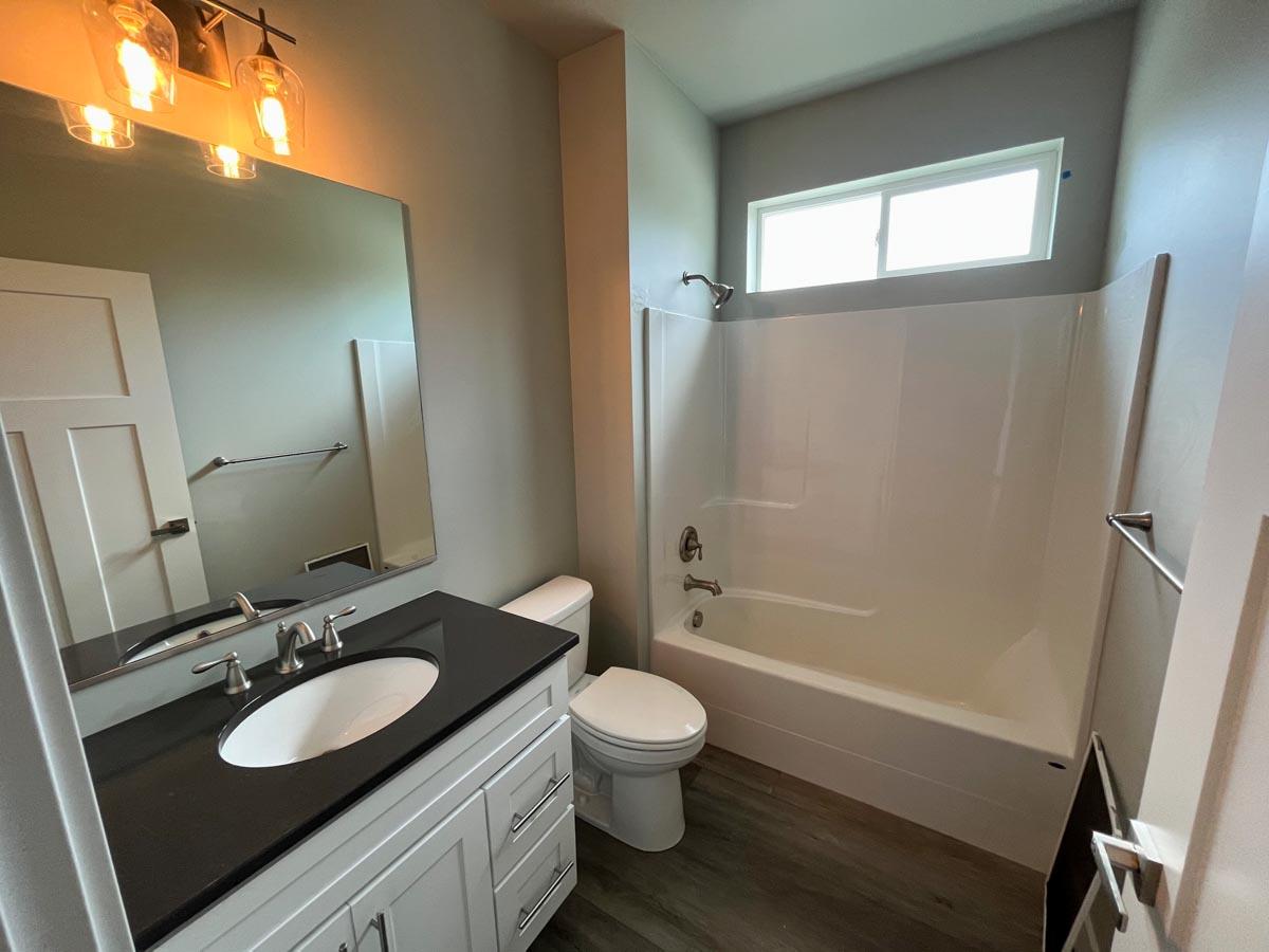 Bathroom with white vanity, dark countertop, toilet, and bathtub with shower, under a horizontal window.