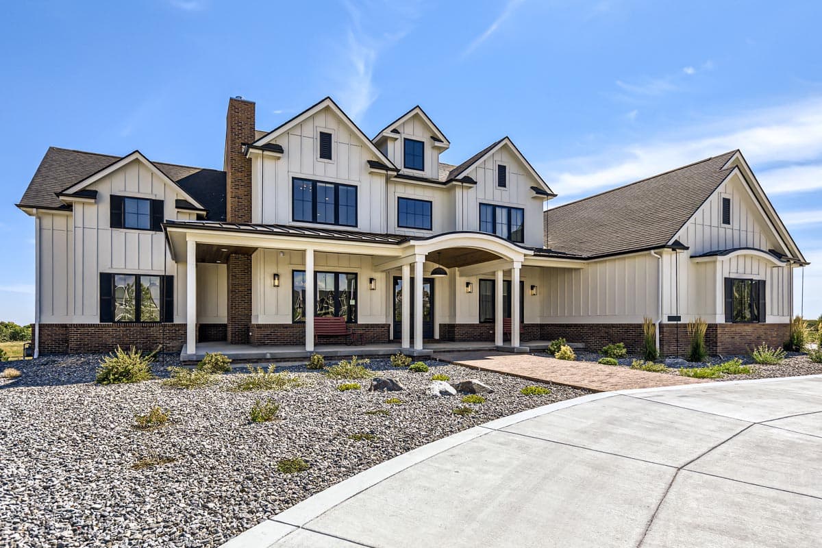 House plan exterior: Modern Farmhouse style, two stories, white vertical siding, brick base, gabled dormers, covered front porch with arched entrance.