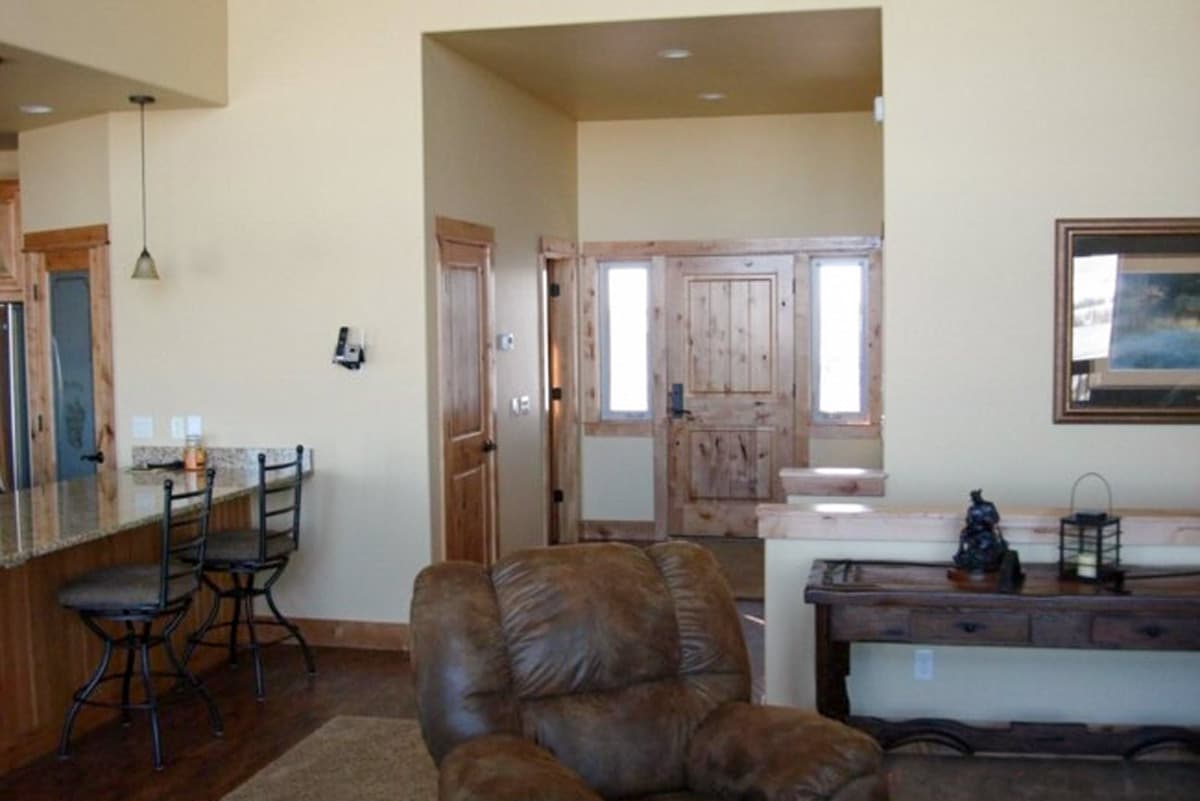 Interior view showing a kitchen island with bar stools, a wooden front door with sidelights, and a brown leather recliner.
