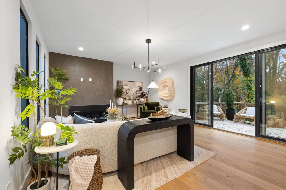 A modern living room with a large window and sliding glass doors that open to a deck. A dark wooden table sits in front of a textured sofa.