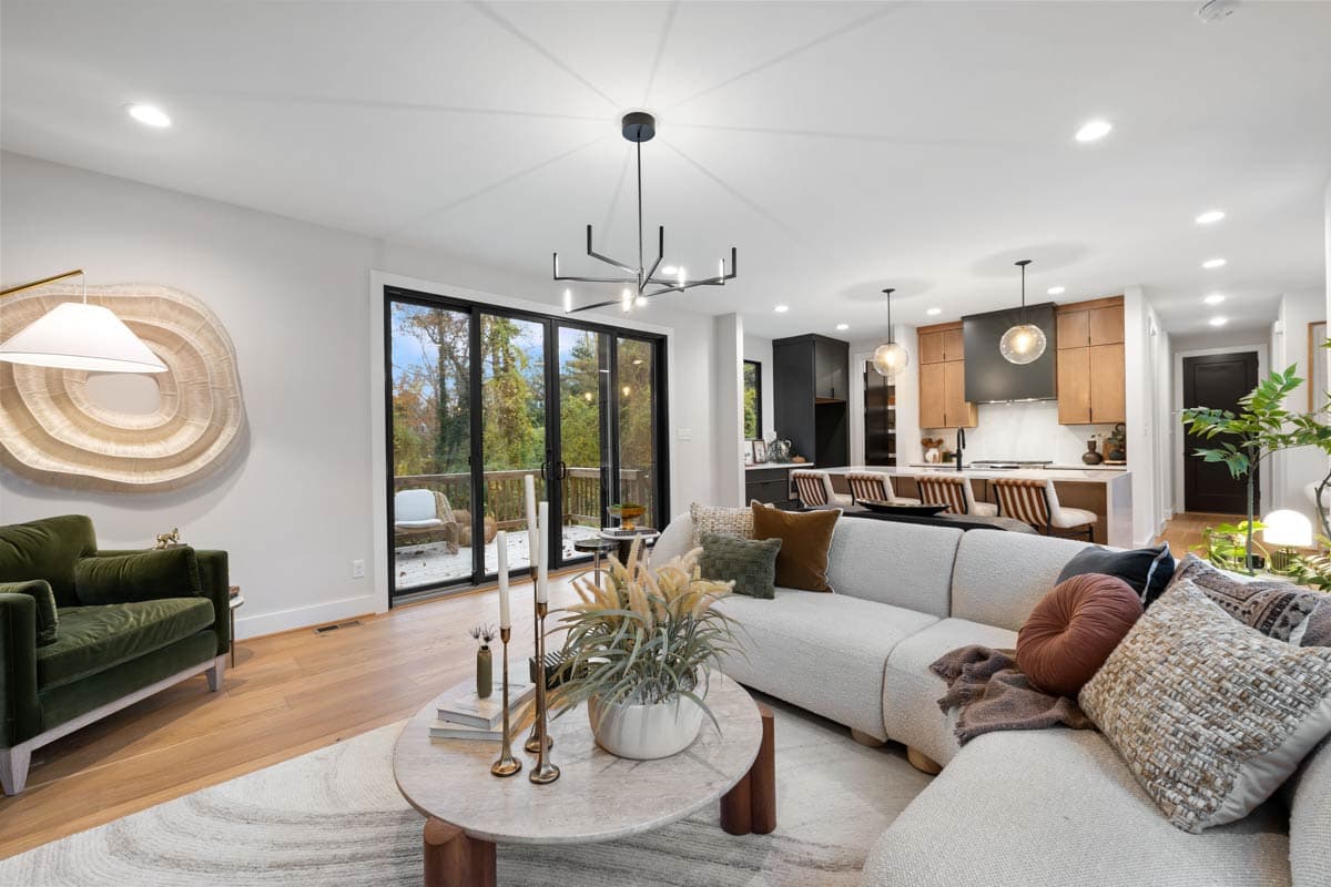 A modern living room with an open floor plan features a large sectional sofa, a round coffee table, and access to a deck overlooking a forest.