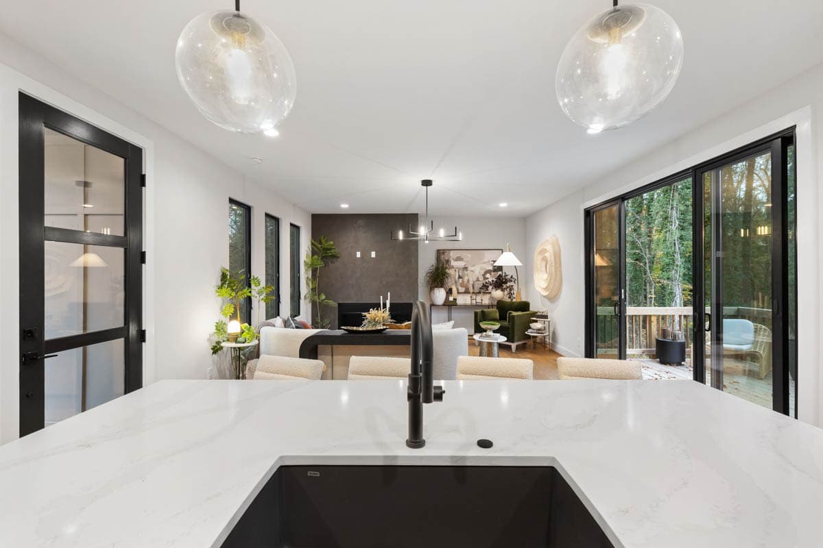 Modern kitchen with marble countertops, leading to a living room with a fireplace and large windows overlooking a deck and trees.