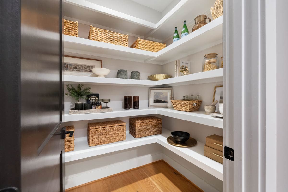 A pantry with white shelves filled with woven baskets, jars, and framed artwork. The pantry features a black door and light wood flooring.