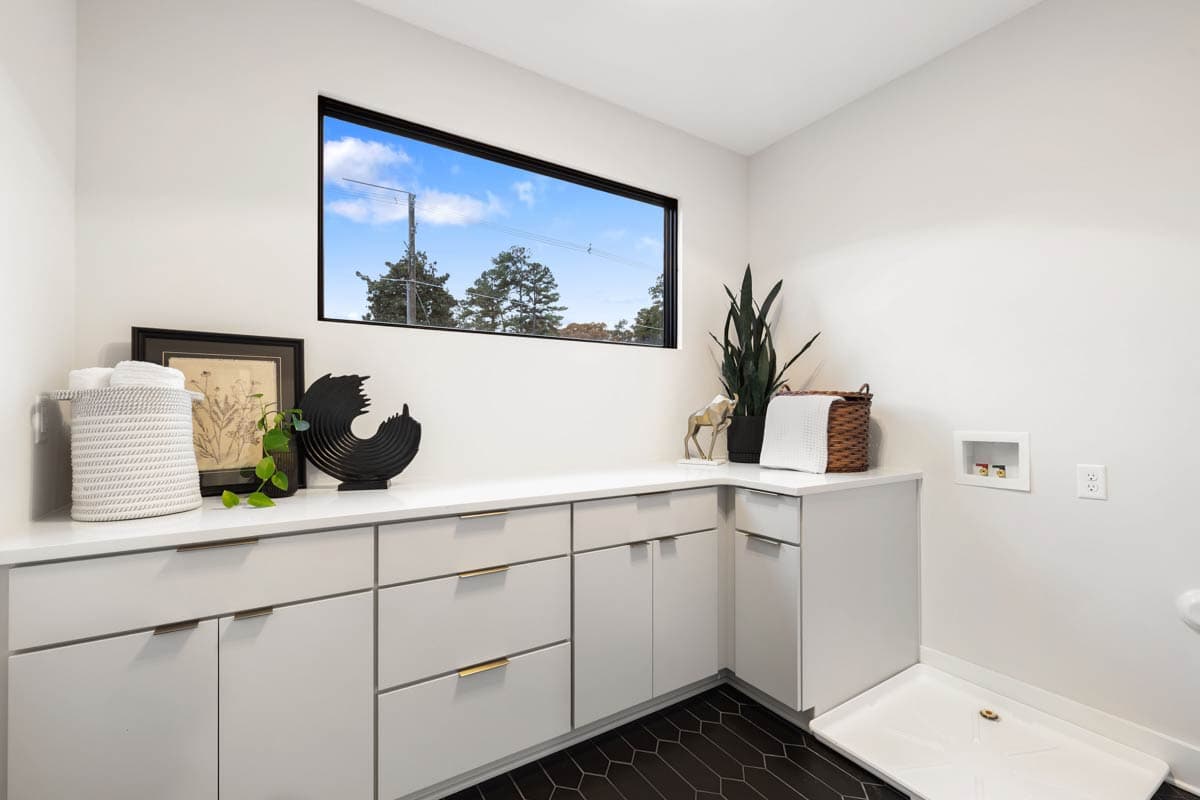 A modern laundry room features white cabinets, a large window showcasing a blue sky, and decorative accents like plants and artwork.