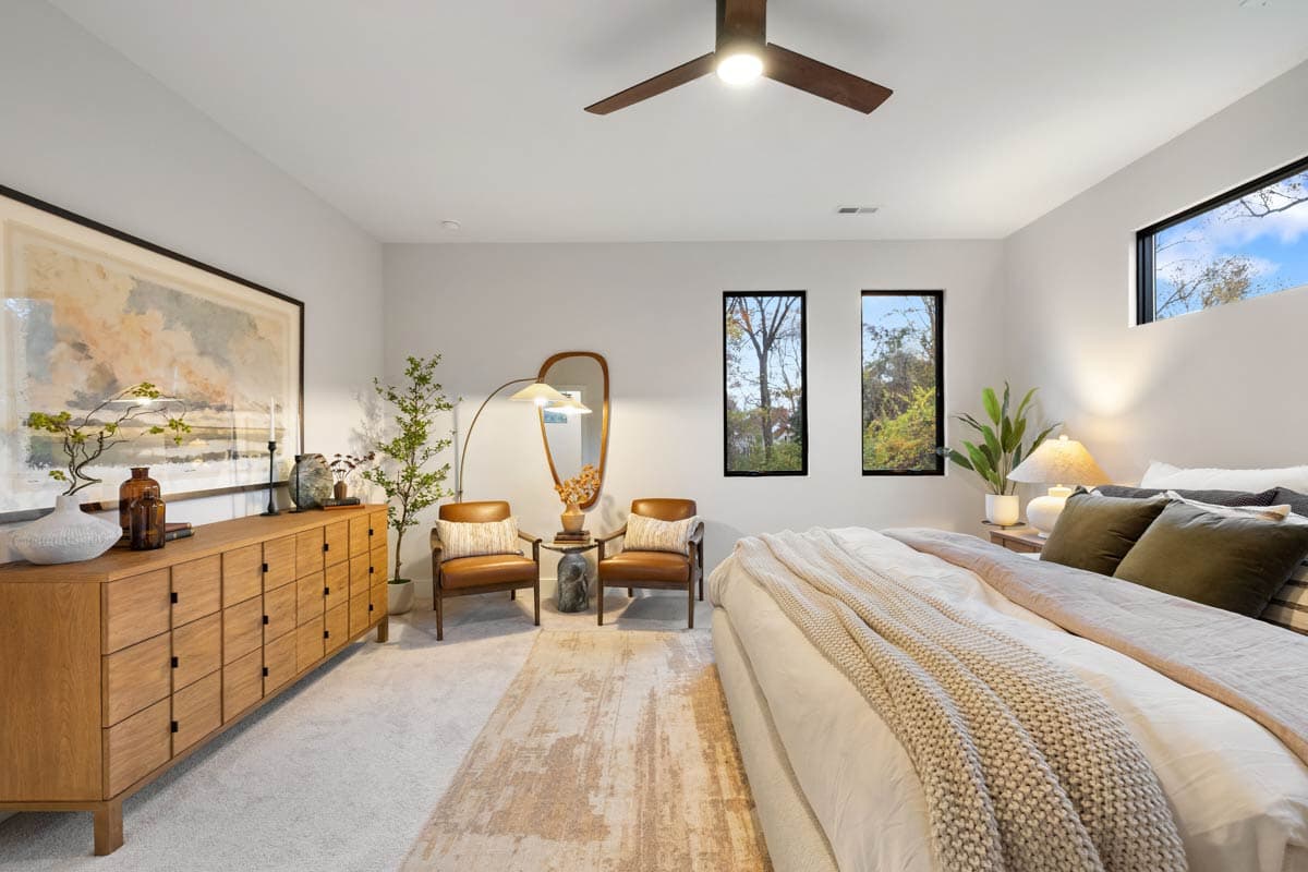 A modern bedroom with a large bed, two leather chairs, and a wooden dresser. Two small windows reveal trees, creating a serene ambiance.