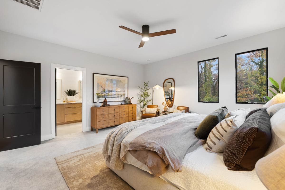 A modern bedroom with a large bed, brown accents, and a view of trees from the windows. A bathroom is seen through an open door.